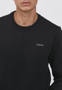 Felpa nera con scollatura rotonda, caratterizzata da un logo sottile sul lato sinistro del petto e dettagli di cucitura testurizzati lungo il colletto.