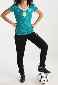 Teal Adidas-paita valkoisella reunanauhoituksella, geometrinen kuvio; mustat housut; mustat jalkapallokengät; pelaaja seisoo jalka mustavalkoisen jalkapallon päällä.