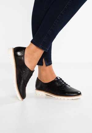 Schwarze glänzende Schnürschuhe mit weißen Sohlen, getragen mit dunkelblauen Slim-Fit-Hosen auf einem weißen Boden.