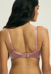 Kobieta z długimi ciemnymi włosami, odwrócona plecami, ubrana w fiołkowy biustonosz z regulowanymi ramiączkami i zapięciem na plecach na hak i oczko, na jednolitym tle.