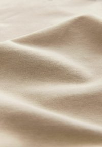 Tissu en coton beige avec une texture douce et lisse, présentant des motifs de stretch et de tissage subtils qui créent de légères ondulations dans le matériau.
