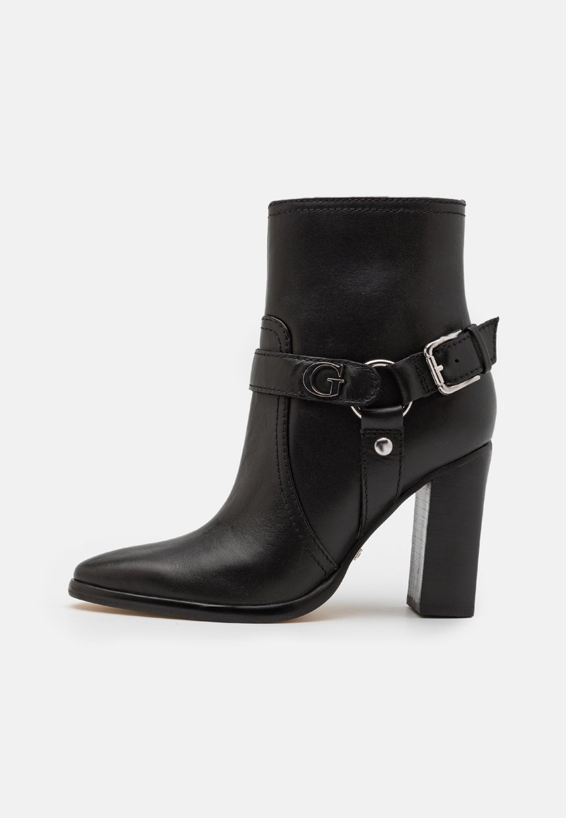 Bottine en cuir noir avec bout pointu, talon de largeur moyenne, bride ajustable à boucle avec un logo décoratif, et texture lisse.