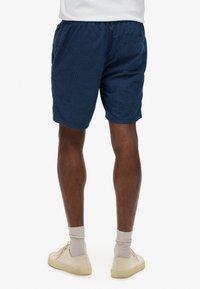 Pantalones cortos azul marino con un patrón texturizado, cinturilla elástica y bolsillos laterales. Llevados con zapatillas crema y calcetines claros.