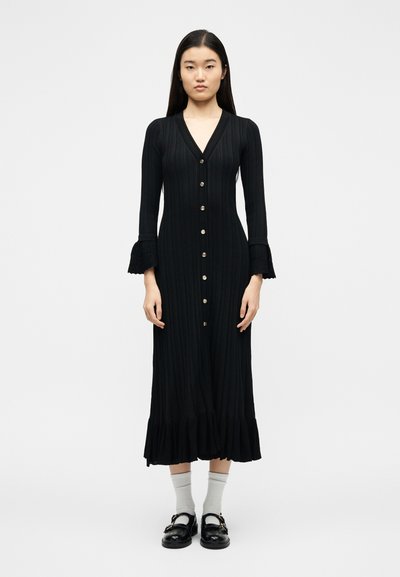 Γυναίκα που στέκεται φορώντας μακρύ μαύρο φόρεμα με κουμπιά και δαντελένια μανσέτα, λευκές κάλτσες και μαύρα παπούτσια με αγκράφα, μπροστά σε απλό λευκό φόντο.