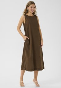Robe sans manches en lin marron avec encolure ronde, poches latérales et silhouette droite, atteignant le milieu du mollet. Sandales de couleur claire.