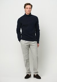 Dunkelblauer gerippter Rollkragenpullover kombiniert mit hellgrauen Hosen. Das Modell trägt dunkelbraune Slipper. Schlichtes, tailliertes Design mit einer klaren Silhouette.