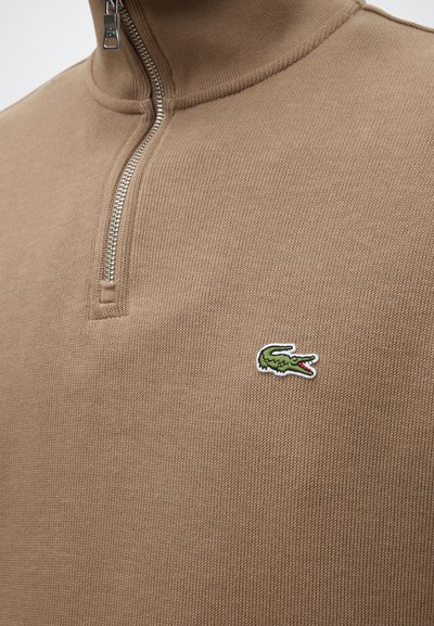 Gros plan d’un pull en maille beige avec une fermeture éclair au quart et un petit logo crocodile vert sur la poitrine.