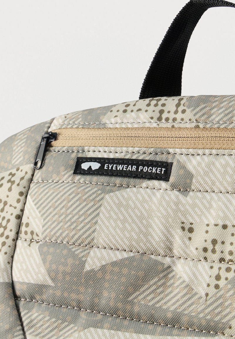 Gros plan sur un coin d'un sac à dos à motif camouflage, avec une petite poche zippée étiquetée « Poche Lunettes » et une boucle de poignée noire au-dessus.