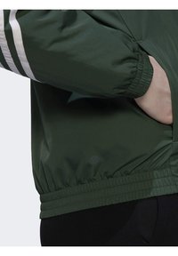 Vihreä takki, jossa valkoiset raidat hihoissa, joustavat hihansuut ja helma. Sileä pinta, sivutaskulla.