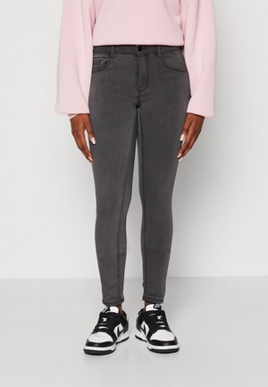 Personne portant un jean gris foncé ajusté, un haut à manches longues rose pâle et des baskets noires et blanches sur un fond uni et clair.