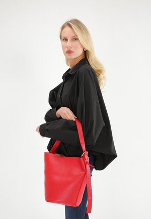 Sac seau en cuir rouge avec une finition texturée, doté d'une courte anse et d'une longue anse ajustable, porté par un modèle en chemise noire.