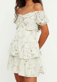 Weißes Blumenkleid mit schulterfreien Puffärmeln, Rüschendetails und gestuftem Design. Das Kleid zeichnet sich durch einen leichten, luftigen Stoff und dezente Erdtöne aus.