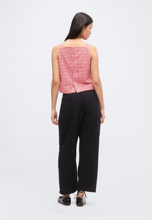 Kobieta w czerwono-białym kraciastym bezrękawniku z guzikami z tyłu, czarnych spodniach z szerokimi nogawkami i czarnych sznurowanych butach, stojąca tyłem.
