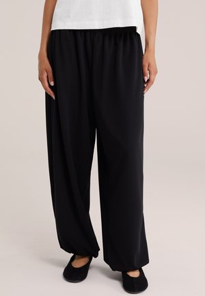 Person trägt weite schwarze Hose, schwarze flache Schuhe und ein weißes Oberteil, steht vor hellem Hintergrund.