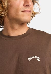 Brun bomulds T-shirt med rund hals og et struktureret hvidt broderet logo på venstre brystområde. Glat overflade.