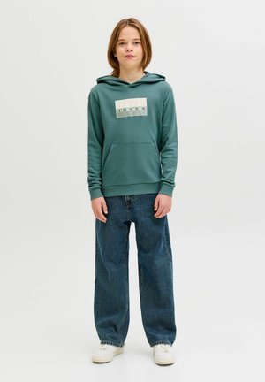 Mladić stoji, nosi zelenu Jack & Jones hoodicu, široke plave traperice i bijele tenisice na jednobojnoj svijetloj pozadini.