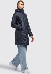Chaqueta impermeable navy con capucha ajustada, cierre frontal y bolsillos laterales, combinada con jeans de pierna ancha de color azul claro y zapatillas blancas.