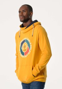 Sweatshirt à capuche jaune avec une poche avant ; présente un graphique circulaire avec des éléments verts, rouges et noirs ; cordon de serrage ajustable.