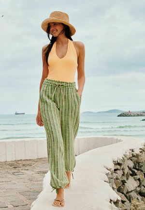 Žena v slamennom klobúku, s žltým tielkom bez rukávov, zelených pruhovaných nohaviciach a sandáloch kráča po zakrivenej pobrežnej ceste s oceánom a loďou v pozadí.