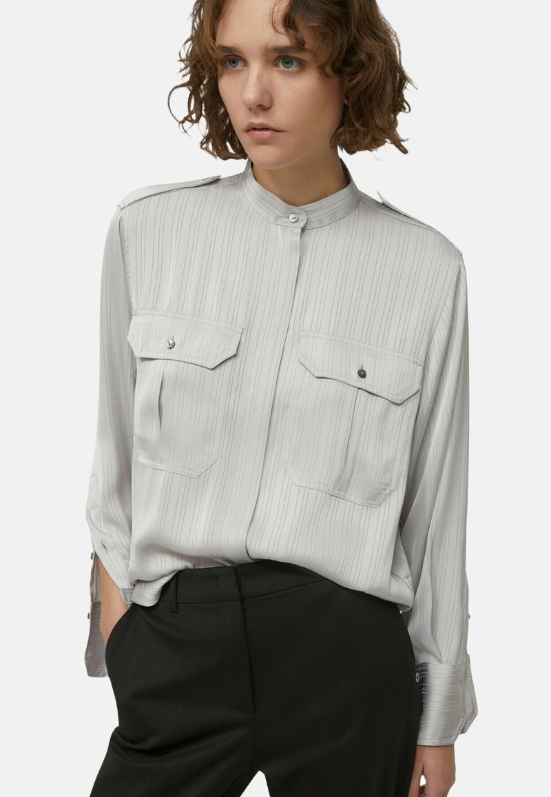 Vrouw in een lichtgrijze gestreepte blouse met knopen en borstzakken, met zwarte broek, poseert met handen in de zakken tegen een witte achtergrond.