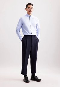 Lichtblauw overhemd met knopen en een gestructureerd patroon, gecombineerd met donkerblauwe geplooide broek en zwarte nette schoenen, handen in de zakken.