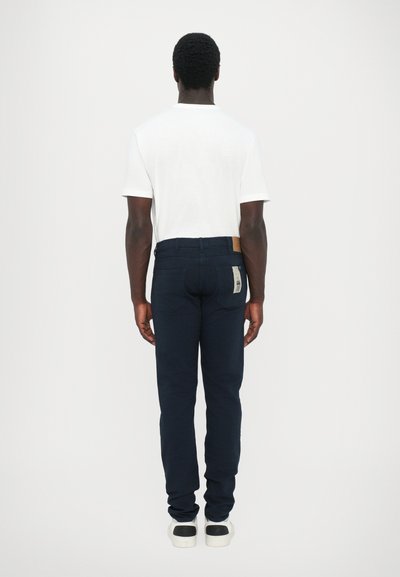 Jeans azul marino de corte slim, material de algodón, etiqueta en la parte trasera y bolsillos en la parte posterior; combinados con una camiseta blanca básica y zapatillas.