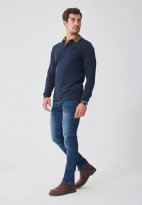 Námořnicky modré polo tričko s dlouhým rukávem a hnědým límečkem, doplněné modrými džínami a hnědými botami. Na hrudi je malé logo.