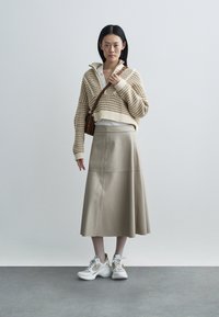 Bézs bőrből készült A-vonalú szoknya, krémszínű, texturált pulóverrel és fehér tornacipővel párosítva. A modell világosszürke háttér előtt áll.