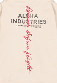 Sudadera de color beige claro con un logo texturizado en negro "ALPHA INDUSTRIES" y un texto estilizado en rojo "Prepare Before Flight" en vertical.