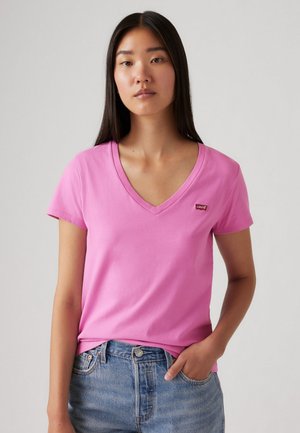 Femme aux longs cheveux foncés portant un t-shirt V rose avec un petit logo Levi's, associé à un jean en denim bleu, une main dans la poche.