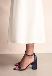 Nohy v čiernych sandáloch na podošve s remienkom okolo členku, v kombinácii s dlhou bielou sukňou, stoja na hladkej béžovej podlahe na bledom pozadí.