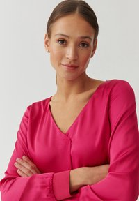 Blusa vibrante rosa con escote en V, mangas largas y puños fruncidos. Textura suave con un ligero brillo, con un diseño de corte cómodo.