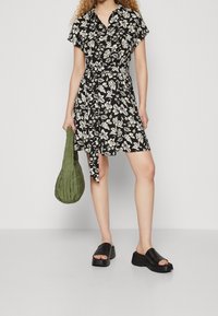 Svart blommig klänning med korta ärmar och knytning i midjan, kombinerad med en grön, vadderad väska och svarta kilklackssandaler med tjock sula.