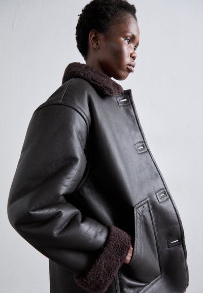 Crna kožna bomber jakna s teksturiranim smeđim umjetnim krznom na ovratniku i manžetama, s bočnim džepovima i čvrstim kopčama na gumbe.