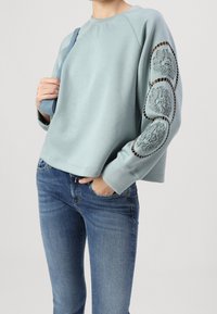 Sweatshirt bleu clair avec motifs de lion texturés sur les manches, associé à un jean bleu. Présente une coupe décontractée et des manches raglan.