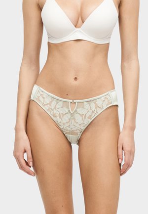 Femme portant un soutien-gorge blanc rembourré et une culotte en dentelle florale assortie, debout devant un fond gris clair uni.