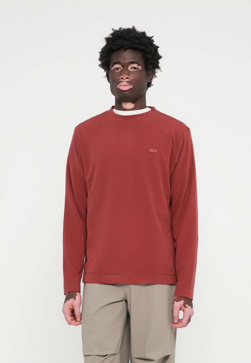 T-shirt à manches longues rouge en tissu doux, avec un col rond et un petit détail d'étiquette sur le devant, associé à un pantalon beige clair.