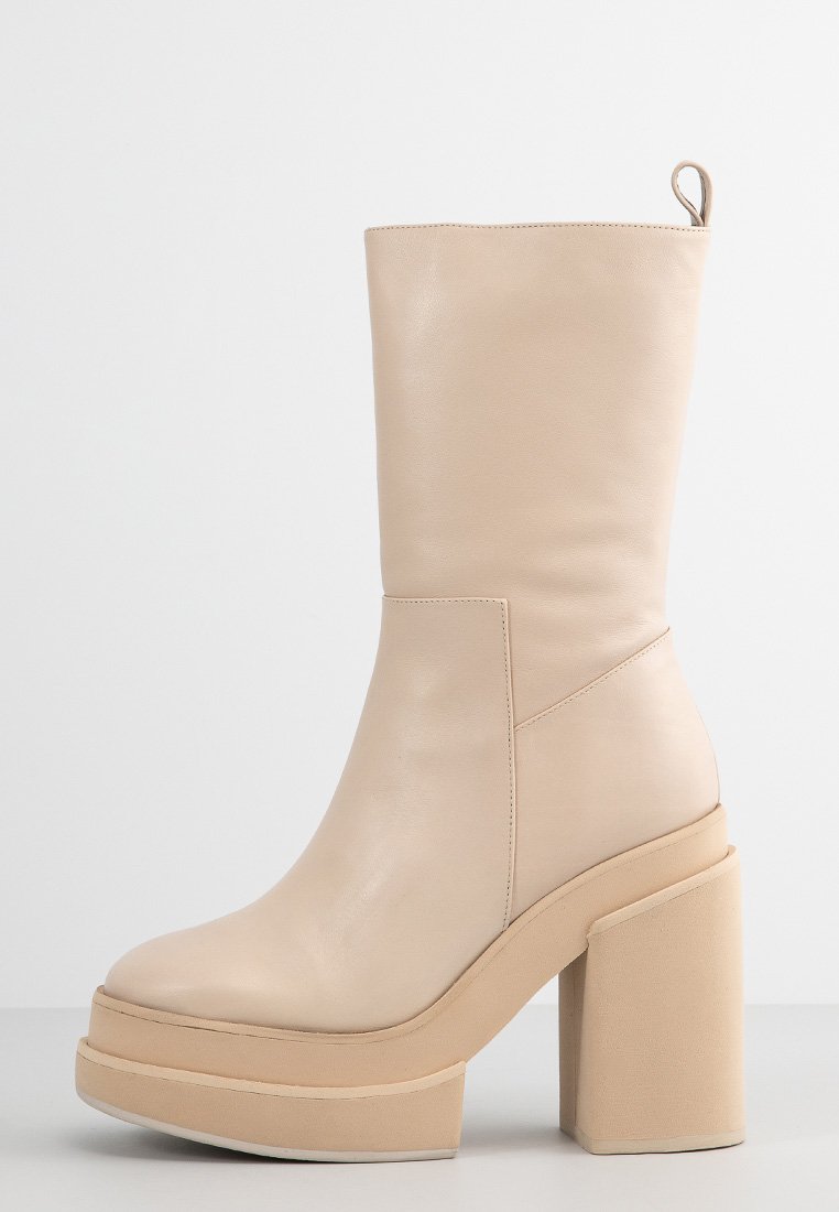 Botín de mujer en tono beige, con un acabado de cuero suave, forma rectangular, tacón alto tipo bloque y suela con plataforma. Presenta un asa de tira para facilitar su colocación.