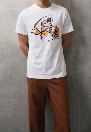 Person trägt weißes T-Shirt mit abstraktem orange-rosa Motiv, kombiniert mit braunen Hosen, steht vor einer grauen Wand.