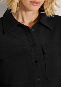 Blonde Frau in einem schwarzen, geknöpften Hemd mit zwei Brusttaschen und einem spitzen Kragen, von den Schultern bis zu den Lippen gezeigt.