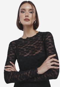 Zwarte kanten top met lange mouwen, voorzien van een gestructureerd patroon, een ronde halslijn en een strakke pasvorm. Model heeft de armen gekruist, waardoor het ontwerp van het kledingstuk goed zichtbaar is.