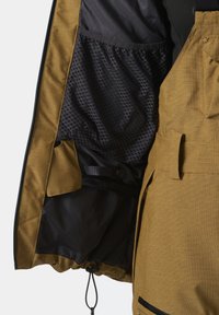 Chaqueta de exterior color marrón dorado con un exterior texturizado, forro de malla negra y un bolsillo interior. Cordon ajustable en el dobladillo.