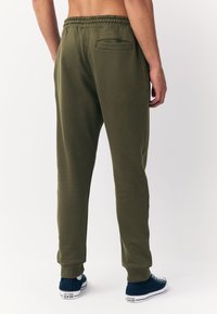 Alyvuogių žali spalvos sportinės kelnės su elastine juosmens dalimi, surišamais kulkšnų kraštais ir galiniu kišene, derinamos su mėlynos spalvos drobiniais sportbačiais.