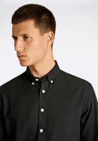 Camicia nera con bottoni, realizzata in tessuto testurizzato, caratterizzata da un colletto classico e bottoni di colore chiaro disposti lungo il davanti.