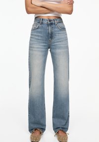 Vaqueros de mezclilla de tiro alto, azul claro, con un corte relajado, lavado desgastado, cinco bolsillos y cierre clásico de botón. Los bordes están bien cortados.