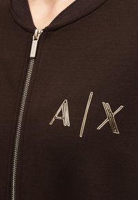 Vêtement marron foncé zippé avec logo métallique doré « A | X » sur la poitrine et une fermeture éclair argentée près du col.