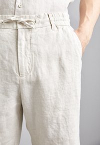 Pantaloni in lino beige chiaro con vita regolabile, tasche laterali e un tessuto testurizzato dall'aspetto naturale e rilassato.
