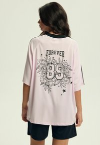 Różowa siatkowa koszulka z dużym czarnym wzorem kwiatowym i napisem "FOREVER" nad numerem "89". Krótkie rękawy i luźny krój. Czarne szorty.