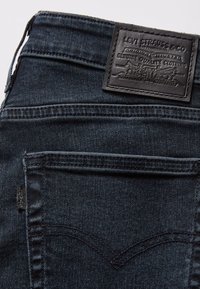 Poche arrière d'un jean Levi's bleu foncé montrant un design arquée cousu, petite étiquette en tissu Levi's et patch en cuir noir avec logo.