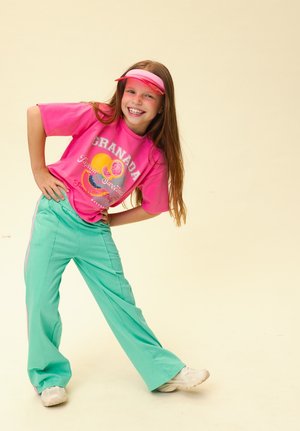Smiling girl wearing a pink visor, pink graphic t-shirt, and teal pants poses with hands on hips against a plain beige background.